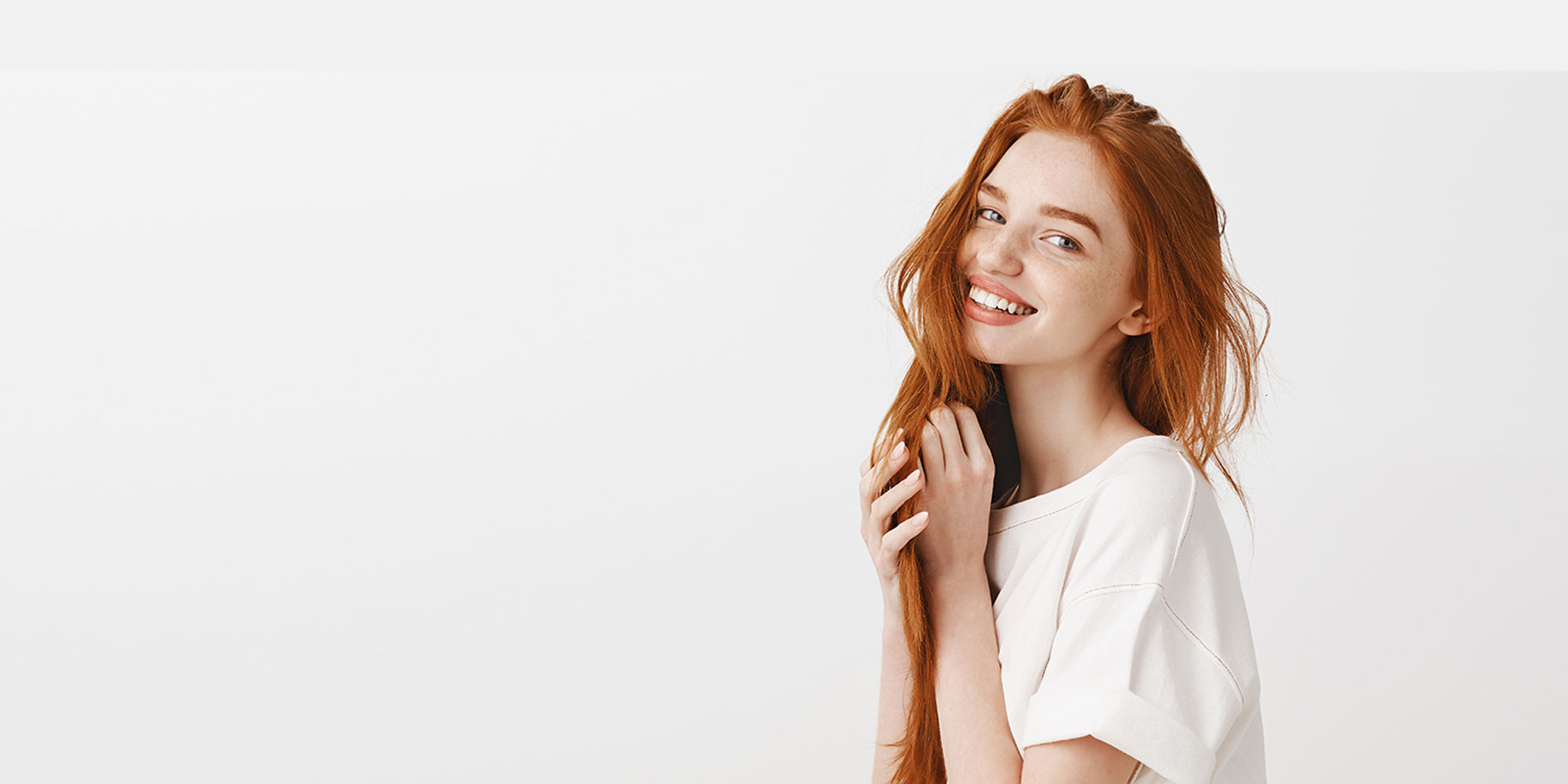 Charming and playful redhead female with long beautiful hair and natural color, smiling tenderly at camera, playing with strands, posing over gray wall